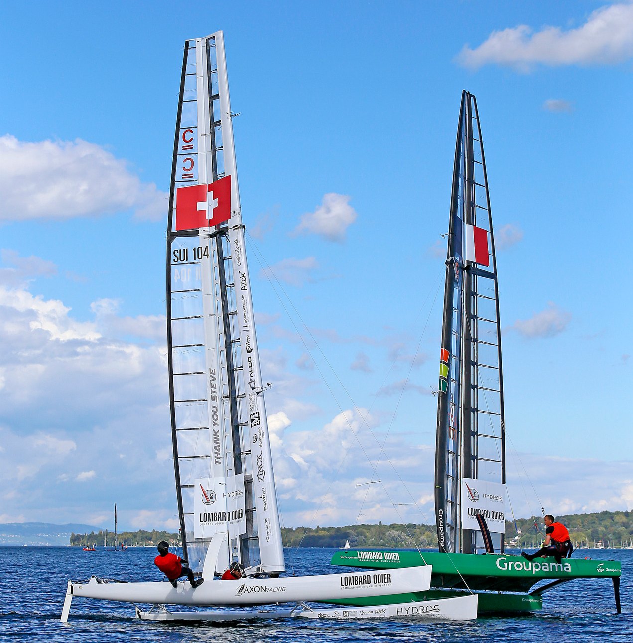 AXON Racing's C-Class catamaran SUI 104 under its wing sail, Swiss flag at the top, racing the green Groupama boat